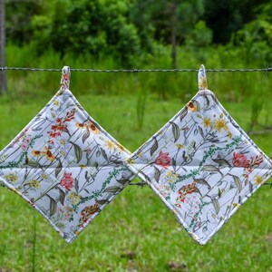 May include: Two square quilted pot holders featuring a white background with a colorful botanical print of wildflowers, including red tulips, yellow daisies, and green foliage. Each pot holder has a fabric loop at the top corner for hanging.