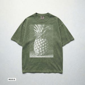 May include: Moss green t-shirt featuring a white dotted pineapple design. The short-sleeved shirt has a relaxed fit and a crew neck. The word "MOSS" is printed at the bottom of the image.