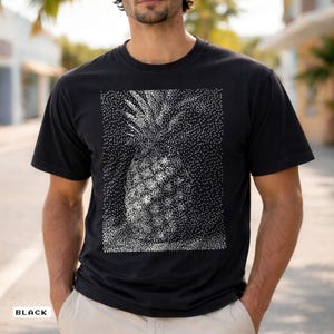 May include: Black t-shirt featuring a detailed, pixelated pineapple design in shades of white and gray. The shirt has a classic crew neck and short sleeves. The word "BLACK" is printed at the bottom left corner of the shirt.