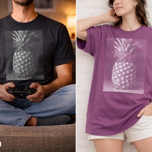 May include: Black and berry-colored t-shirts featuring a white dotted pineapple design. The shirts are short-sleeved and have a relaxed fit. The black shirt is worn by a person holding a game controller. The berry shirt is worn by a person with their hand in their pocket.