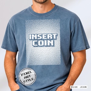 Puede incluir: Camiseta azul vaquero con el texto "INSERT COIN" en un diseño pixelado. La camiseta tiene un logotipo redondo de estilo pixel art en la parte inferior izquierda. La camiseta está hecha de un material suave.