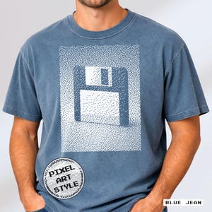 Könnte beinhalten: Blaues Jeans-T-Shirt mit Pixel-Art-Design einer Diskette. Das Design ist weiß und vor einem gepunkteten Hintergrund. Das T-Shirt hat einen Rundhalsausschnitt und kurze Ärmel. Ein kreisförmiges Logo mit dem Text "PIXEL ART STYLE" befindet sich unten links.