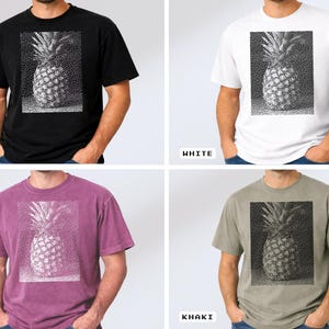 May include: A black, white, berry, and khaki t-shirt featuring a detailed pineapple graphic. The pineapple design is in a rectangular frame. The shirts are short-sleeved and made of a soft material.