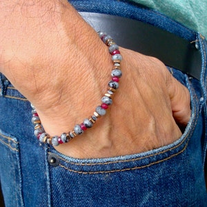 May include: A bracelet with gray and red beads and silver accents. The bracelet is on a person's wrist.
