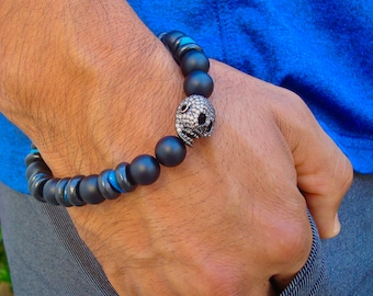 May include: A black beaded bracelet with a skull charm. The skull is encrusted with small, clear rhinestones. The bracelet has blue accents.