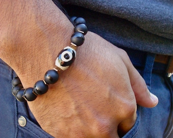 May include: A black beaded bracelet with a gold-toned evil eye charm. The bracelet is worn on a person's wrist.
