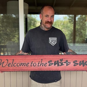 May include: A red wooden sign with the phrase "Welcome to the Shit Show" in black lettering. The sign has a decorative edge and is held by a person wearing a black t-shirt with a "Thrashin USA" logo.