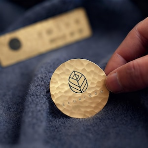 May include: A gold-colored, hammered metal disc with the text "ECO BLOOM" and a leaf design. The disc is held over a blue fabric. A rectangular tag with a black circle is in the background.