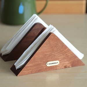 May include: Two wooden napkin holders in a triangular shape, filled with white napkins. The holders are made of dark brown wood and have a small, oval-shaped gold label that reads "SAVORSPICE". The holders are on a light-colored wooden surface.