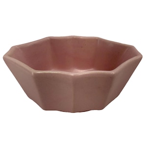 May include: A small, pink ceramic bowl with a geometric, faceted design. The bowl has a wide, open top and a series of angled sides, creating a multi-sided shape. The bowl is a solid, soft pink color.