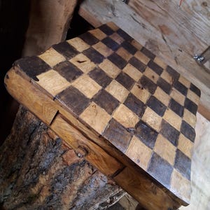 May include: A handcrafted wooden chess board with a checkerboard pattern of light and dark squares. The board is set on a rustic wooden base, showcasing the natural grain and texture of the wood. The chess board is a classic game for all ages.