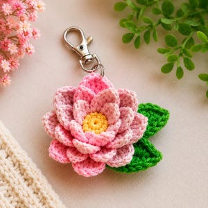 May include: A crochet keychain featuring a multi-layered pink lotus flower with a bright yellow center and two green leaves attached to a silver-toned metal lobster clasp. The flower is crafted with detailed yarn stitching and a textured petal design.