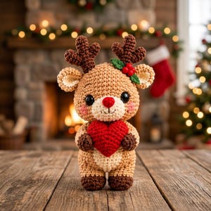 May include: A crocheted reindeer plush toy with light brown yarn, dark brown antlers, and a red nose. The reindeer has large black eyes, pink cheeks, and holds a bright red crocheted heart. A small green leaf and red berry accent sit on one antler.