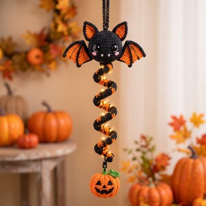 May include: A hanging crochet decoration featuring a black bat with orange inner wings and white fangs, attached to a black and orange spiral tail illuminated by small string lights, ending with a small orange crocheted jack-o'-lantern at the base.