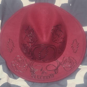 May include: A burgundy fedora-style hat with a wide brim. The hat features a black, glittery design of roses, leaves, a skull, and a lightning bolt. The hat is a stylish accessory.