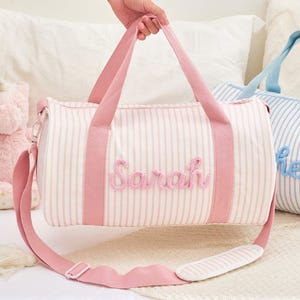 May include: Two striped duffel bags, one pink and one blue, with embroidered names. The pink bag has a pink strap and the name "Sarah". The blue bag has a blue strap and the name "Hel". A pink unicorn plush toy is visible.