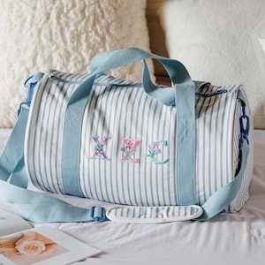 May include: A striped duffel bag with blue accents and embroidered floral initials. The bag features a light blue shoulder strap and handles. The bag is on a white surface with open magazines and pillows in the background.