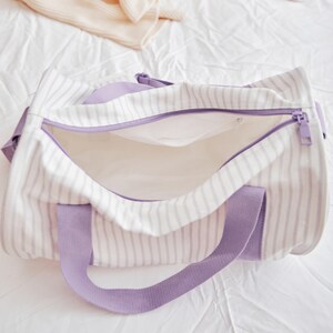 May include: Open, striped canvas tote bag with lavender accents. The bag features a white and gray striped pattern, lavender handles, and a matching zipper. The interior is a solid white color. A folded sweater is in the background.