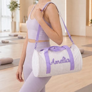 May include: A white and gray striped duffel bag with purple accents, including the shoulder strap and handles. The name "Amelia" is embroidered in purple on the side. The bag is cylindrical and appears to be made of fabric.