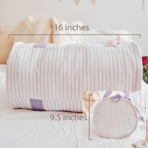 May include: A white and lavender striped bolster pillow, 16 inches long, with a matching round bag, 9.5 inches in diameter. The pillow and bag feature a soft, vertical stripe pattern and are made of a light-colored fabric. The bag has a lavender handle.