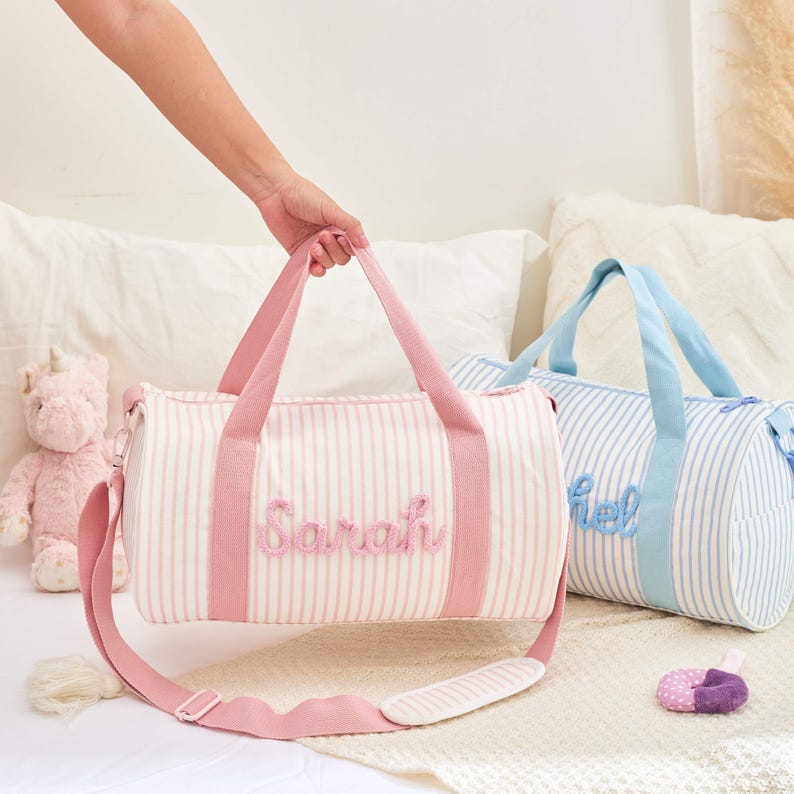 May include: Two striped duffel bags, one pink and one blue, with embroidered names. The pink bag has a pink strap and the name "Sarah". The blue bag has a blue strap and the name "Hel". A pink unicorn plush toy is visible.