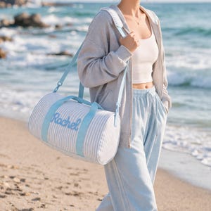 May include: A light blue and white striped duffel bag with the name "Rachel" embroidered in light blue. The bag has light blue handles and a shoulder strap. The person in the image is wearing a gray hoodie, white top, and light blue sweatpants.