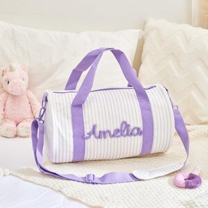 May include: White and lavender striped duffel bag with lavender handles and the name "Amelia" embroidered on the front. The bag has a detachable shoulder strap. A pink unicorn stuffed animal is visible in the background.