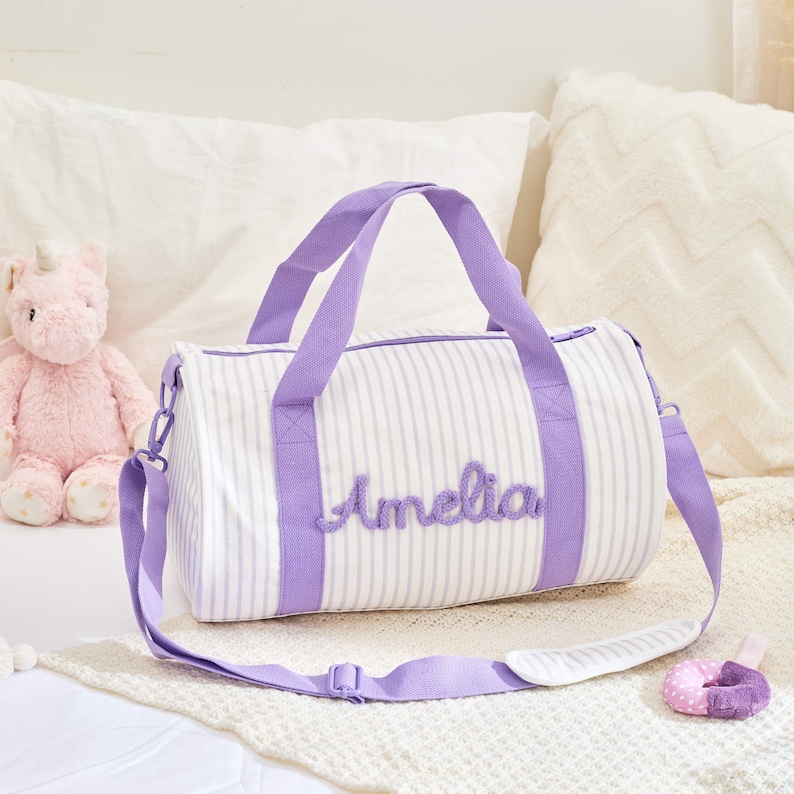May include: White and lavender striped duffel bag with lavender handles and the name "Amelia" embroidered on the front. The bag has a detachable shoulder strap. A pink unicorn stuffed animal is visible in the background.
