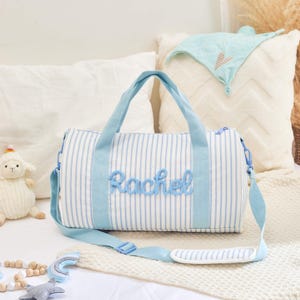 May include: A blue and white striped duffel bag with the name "Rachel" embroidered in blue script. The bag has blue handles and a detachable shoulder strap. A plush lamb and other decorative items are in the background.