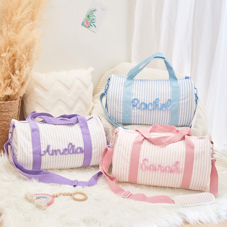 May include: Three personalized duffel bags in pastel colors. Each bag features a white and blue striped pattern with a name embroidered in a matching color. The bags have handles and adjustable shoulder straps. One bag is lavender, one is light blue, and one is pink.