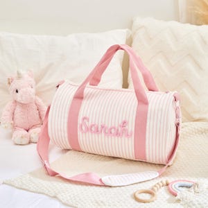 May include: A pink and white striped duffel bag with the name "Sarah" embroidered on the front. The bag has pink handles and a shoulder strap. A pink unicorn plush toy and a wooden rainbow toy are also in the image.
