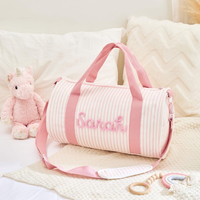 May include: A pink and white striped duffel bag with the name "Sarah" embroidered on the front. The bag has pink handles and a shoulder strap. A pink unicorn plush toy and a wooden rainbow toy are also in the image.