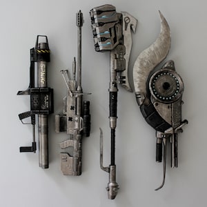 Könnte beinhalten: Vier futuristische Metallwaffen werden vor einer schlichten Wand präsentiert. Die Sammlung umfasst ein zylindrisches Gerät mit dem Text "SPNK", eine gewehrartige Waffe, einen Hammer und eine große, gebogene Klinge mit einem kreisförmigen Bauteil.