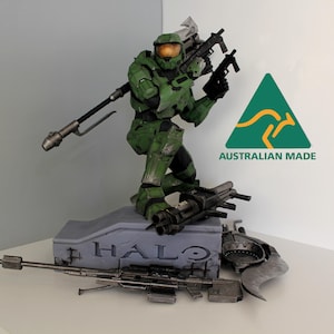 May include: A detailed green and grey Halo action figure of Master Chief holding weapons. The figure is posed on a grey base with the word "HALO" and various weapons. The image also includes an Australian Made logo.
