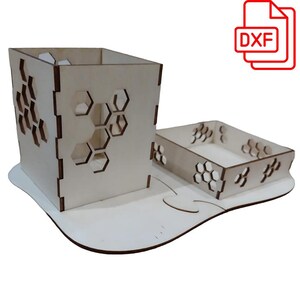 Può includere: Un set di organizzatori da scrivania in legno con un portapenne quadrato e un vassoio rettangolare. Entrambi i pezzi presentano ritagli a nido d'ape e sono realizzati in legno di colore chiaro. Il set poggia su una base bianca di forma organica. Un'icona del file DXF si trova in alto a destra.