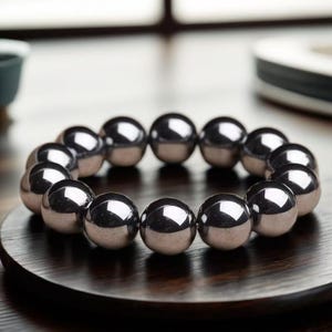 Könnte beinhalten: Ein silberfarbenes Armband aus kugelförmigen Perlen. Die Perlen sind kreisförmig angeordnet, reflektieren das Licht und erzeugen ein glänzendes Aussehen. Das Armband wird auf einer dunklen Holzoberfläche präsentiert.