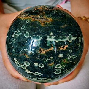 May include: A polished, spherical gemstone with a dark green base color, speckled with white and accented with brown and gold inclusions. The orb is held in two hands, showcasing its smooth, reflective surface. This decorative sphere is a unique home decor item.