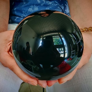 May include: A large, polished black sphere held in two hands. The sphere reflects the surrounding environment, including a room with a window, a person, and other objects. The sphere is smooth and highly reflective.