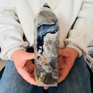 Puede incluir: Una torre de piedra pulida con base hexagonal y punta afilada. La superficie presenta un patrón complejo de colores arremolinados que incluyen tonos negros, gris oscuro, crema y blancos, con pequeñas manchas de inclusiones minerales naranjas.