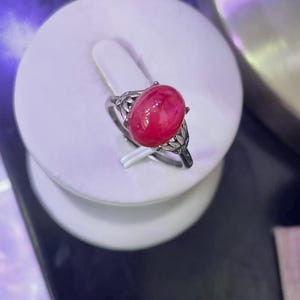 May include: A silver ring featuring an oval, vibrant red gemstone. The ring has an ornate, filigree design on the sides of the band, with small accent stones. The gemstone is the focal point, set prominently in the center of the ring.