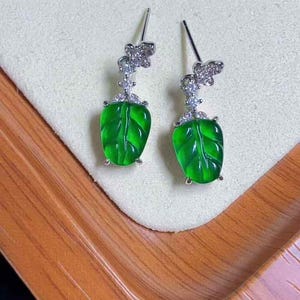 Könnte beinhalten: Silberne Ohrringe mit floralem Design und leuchtend grünen, blattförmigen Anhängern. Die Ohrringe werden auf einer cremefarbenen Oberfläche präsentiert, mit einem Holztablett im Hintergrund.