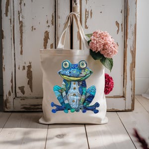 May include: A cream-colored tote bag featuring a whimsical frog design. The frog is primarily blue with patchwork denim details, large eyes, and a smiling expression. The bag has sturdy handles and is set against a rustic, white-painted wooden background.