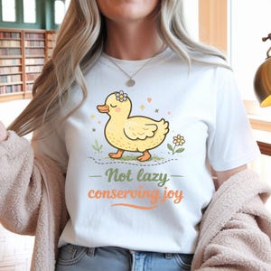 May include: White t-shirt featuring a cartoon yellow duck with a flower crown, walking on a dotted line. The text "Not lazy conserving joy" is printed below the duck in green and orange. The shirt is a casual wear item.
