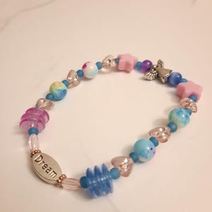 May include: A beaded bracelet with a variety of colorful beads. The bracelet features pink star-shaped beads, blue and purple beads, and a silver charm with the word "Dream". The bracelet is approximately 7 inches in length.