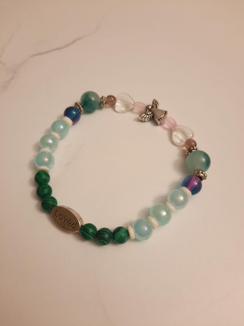 Puede incluir: Una pulsera de cuentas con cuentas verdes, azules y rosas. Incluye un dije de &aacute;ngel plateado y un dije ovalado de metal con la palabra "Loved". Las cuentas var&iacute;an en tama&ntilde;o y textura.