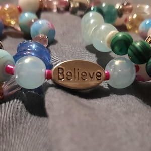Puede incluir: Primer plano de pulseras de cuentas con varias cuentas de colores. Una pulsera tiene la palabra "Dream" en un dije ovalado, otra "Believe" y una tercera "Loved". Las cuentas son en tonos de azul, verde, rosa y blanco.