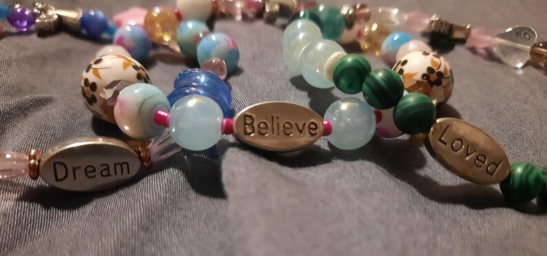 Puede incluir: Primer plano de pulseras de cuentas con varias cuentas de colores. Una pulsera tiene la palabra "Dream" en un dije ovalado, otra "Believe" y una tercera "Loved". Las cuentas son en tonos de azul, verde, rosa y blanco.
