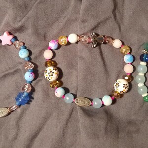 Puede incluir: Tres pulseras de cuentas con varias cuentas de colores, incluyendo estrellas rosas, esferas azules y blancas y dijes decorativos. Una pulsera tiene la palabra "Dream", y otra la palabra "Believe".