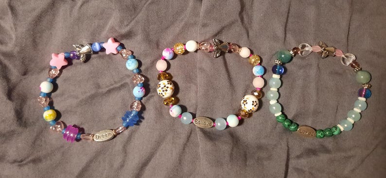 Puede incluir: Tres pulseras de cuentas con varias cuentas de colores, incluyendo estrellas rosas, esferas azules y blancas y dijes decorativos. Una pulsera tiene la palabra "Dream", y otra la palabra "Believe".