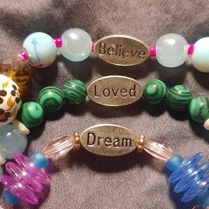 Puede incluir: Tres pulseras de cuentas con palabras inspiradoras. La pulsera superior tiene cuentas azules claras y blancas con la palabra "Believe". La pulsera del medio tiene cuentas verdes con la palabra "Loved". La pulsera inferior tiene cuentas azules y moradas con la palabra "Dream".